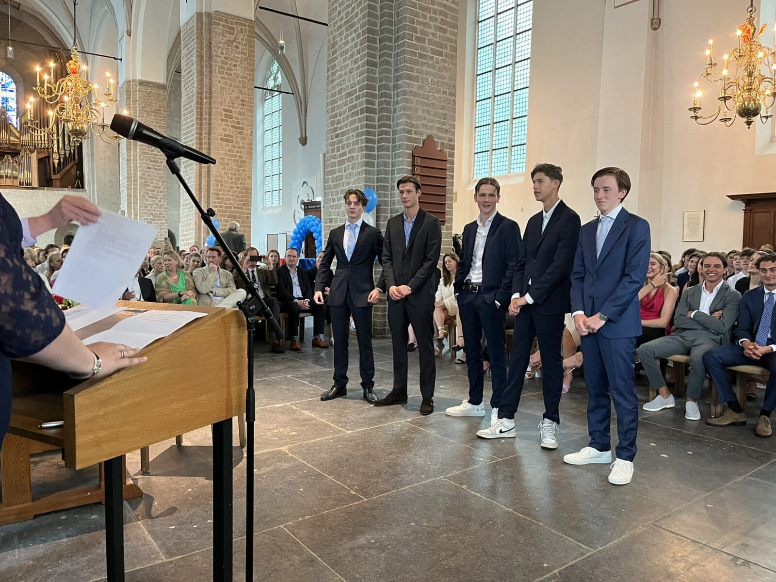 Diploma-uitreiking in een kerk met vijf studenten in pak.