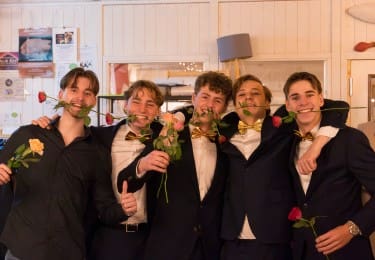 Vijf lachende jonge mannen in pak met rozen in hun mond.