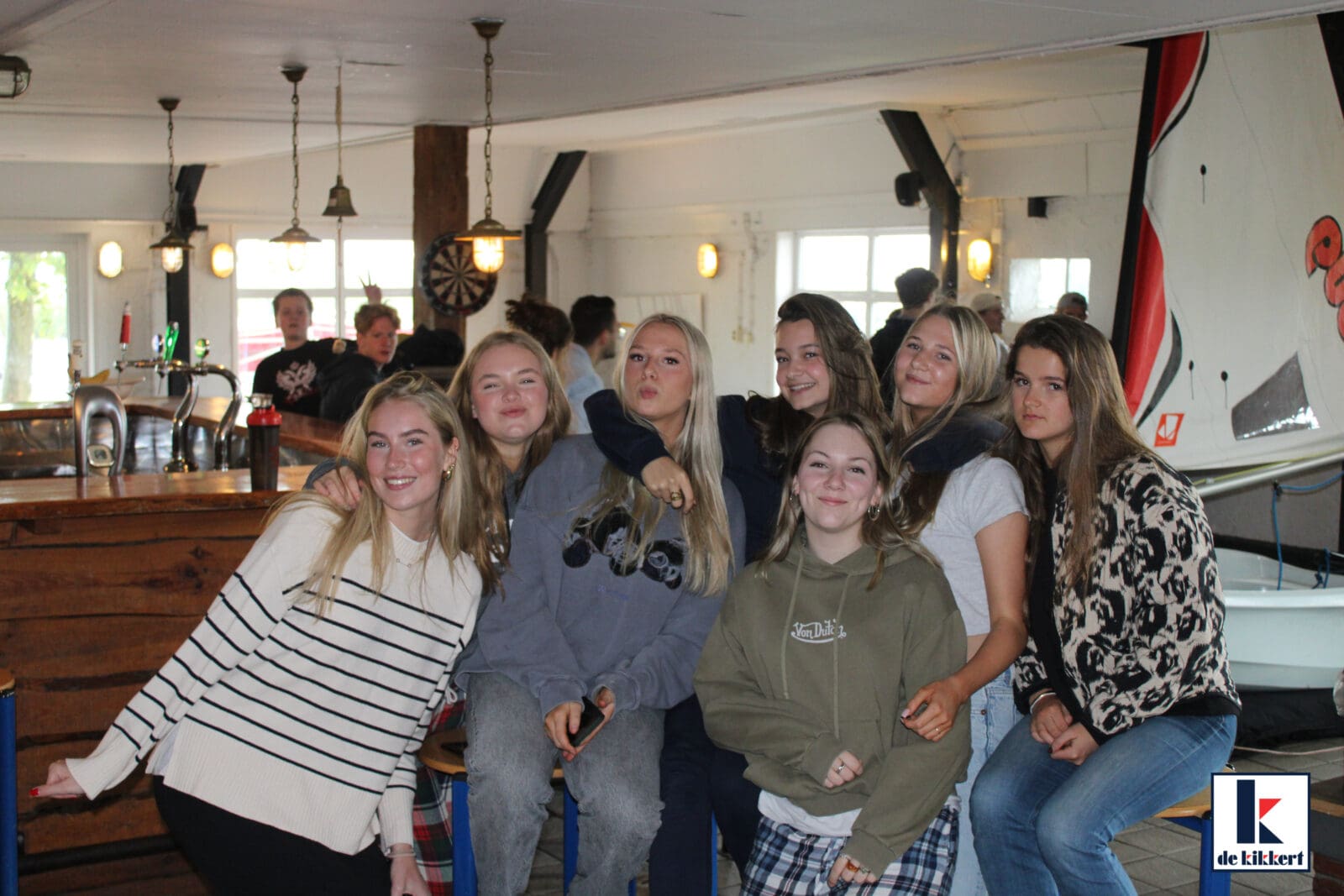 Groep studenten poseren in een café met een boot op de achtergrond.