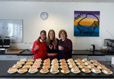 Drie vrouwen poseren met borden vol tosti's in een keuken.