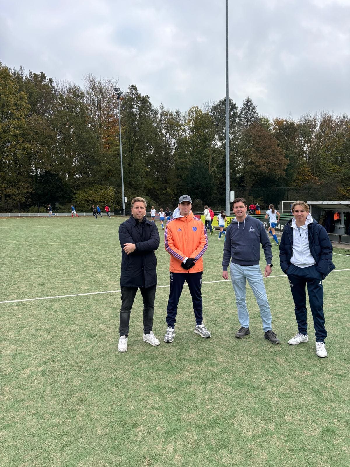 Vier mannen op een hockeyveld met bomen op de achtergrond.